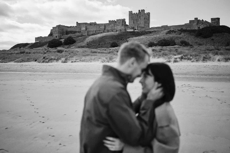 Black and white portrait of an engaged couple embracing on the beach beneath Bamburgh Castle
